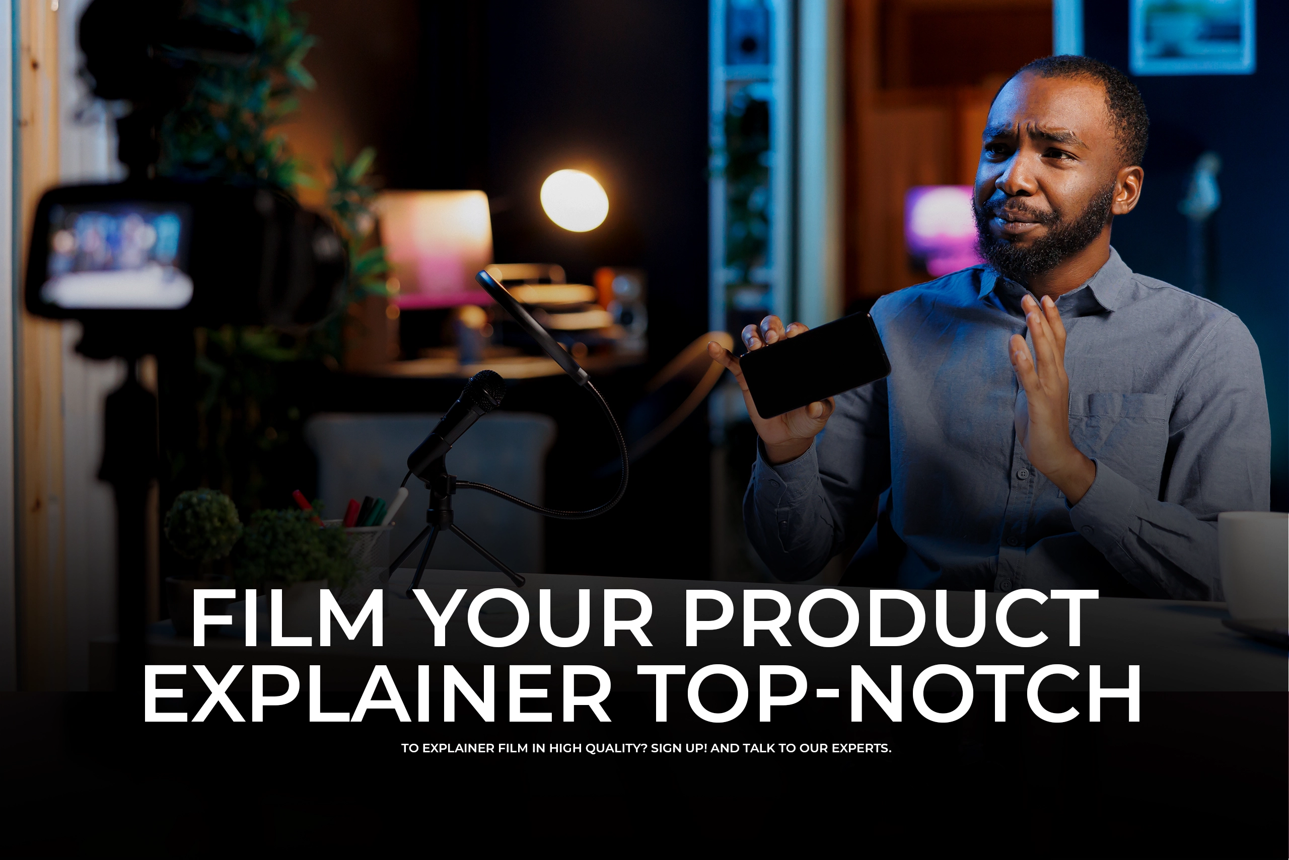 A professional studio scene featuring a man sitting at a desk, holding up a smartphone toward a camera while talking into a microphone. Bold text reads "FILM YOUR PRODUCT EXPLAINER TOP-NOTCH."