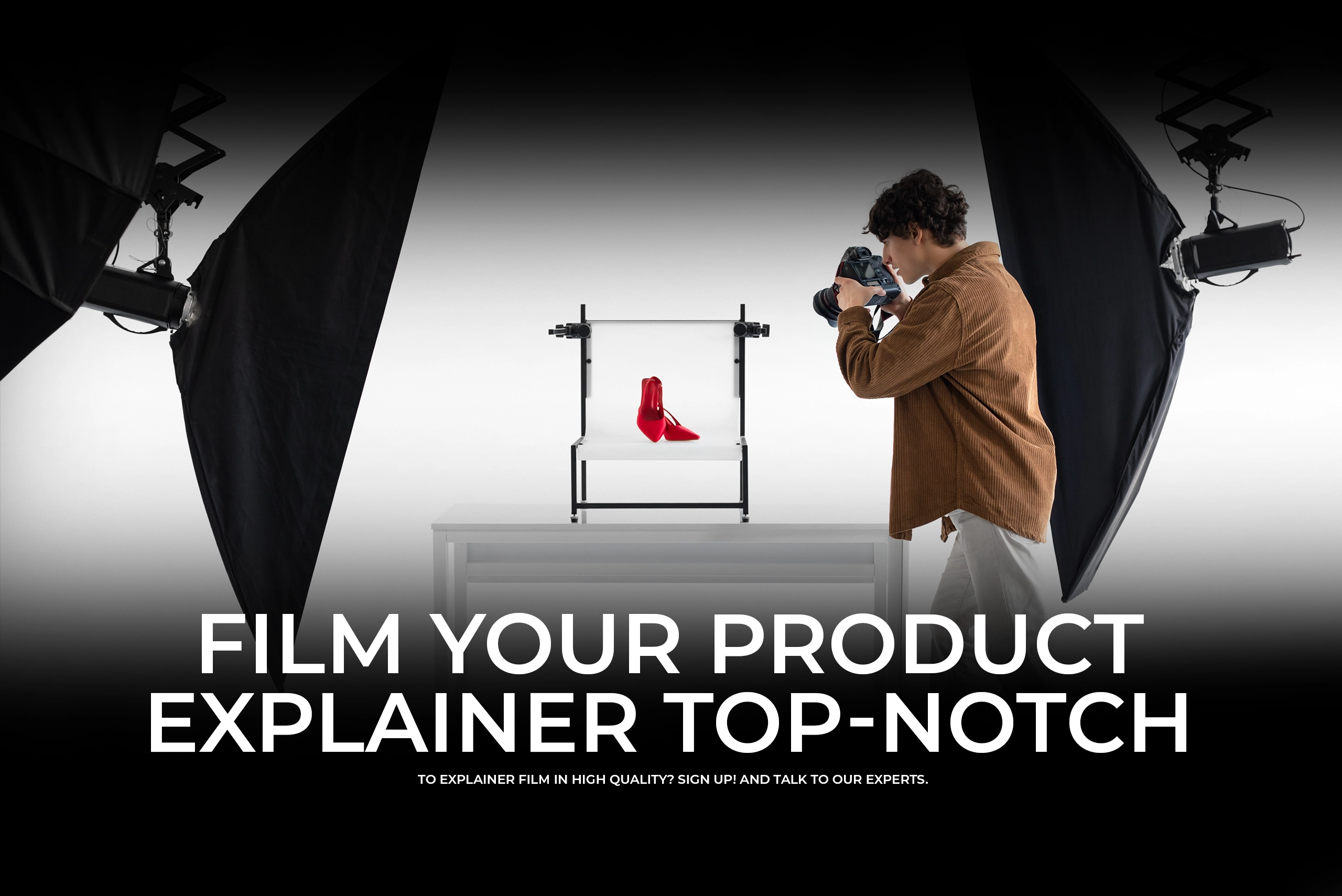 A high-key studio photograph showing a photographer in a brown corduroy shirt taking a picture of a pair of red high-heeled shoes on a white product table, flanked by large studio softboxes. Bold text reads "FILM YOUR PRODUCT EXPLAINER TOP-NOTCH."