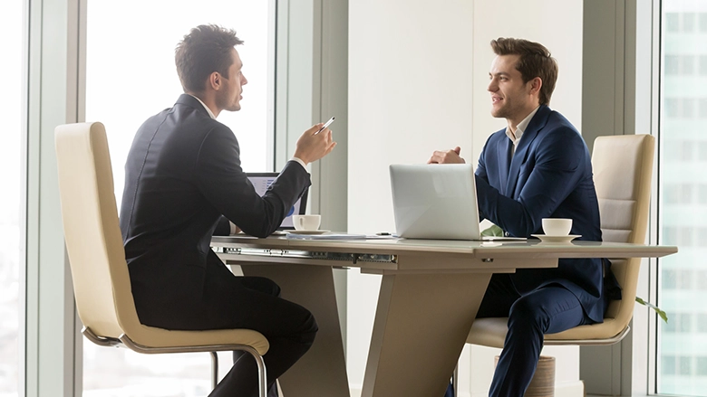 Two men in professional dark suits are seated across from each other at a modern desk in a high-rise office with large windows, engaged in a serious business discussion with laptops and coffee