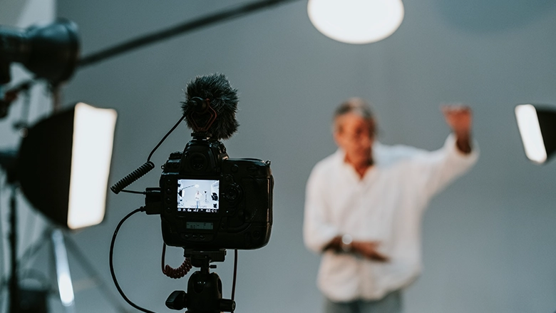 A professional camera on a tripod with a fluffy windshield microphone is focused on a man in a white shirt who is blurred in the background, gesturing during a studio shoot.