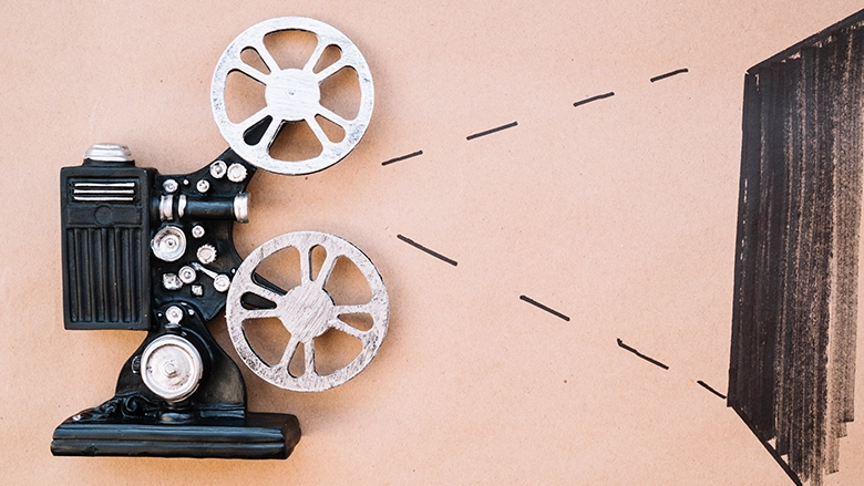 A flat-lay image of a vintage-style black film projector with silver reels, positioned against a tan background with hand-drawn dotted lines indicating light rays projecting onto a dark screen.